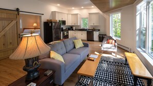 A modern kitchen with white cabinets, a refrigerator, and a stove. There is a gray sofa with yellow pillows, a wooden coffee table, and a patterned rug. - Peaceful, family-friendly Maine getaway at newly built scenic lakefront cottage  (Clifton)
