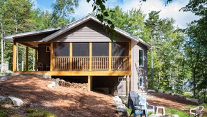 A cabin with a wooden deck, a covered porch, and a view of the surrounding forest. - Peaceful, family-friendly Maine getaway at newly built scenic lakefront cottage  (Clifton)