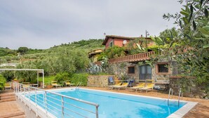 Una piscina con barandilla metálica, rodeada por un patio de ladrillo. Hay sillones de descanso y una pequeña mesa cerca.