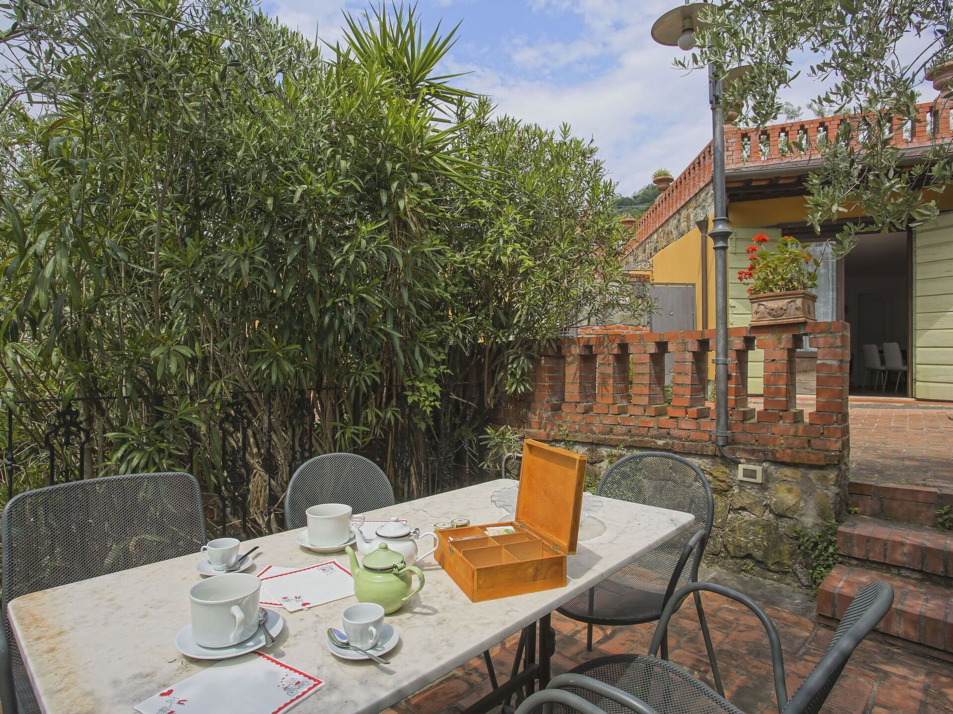 A table set for tea with cups, saucers, and a teapot, surrounded by chairs and a brick wall.