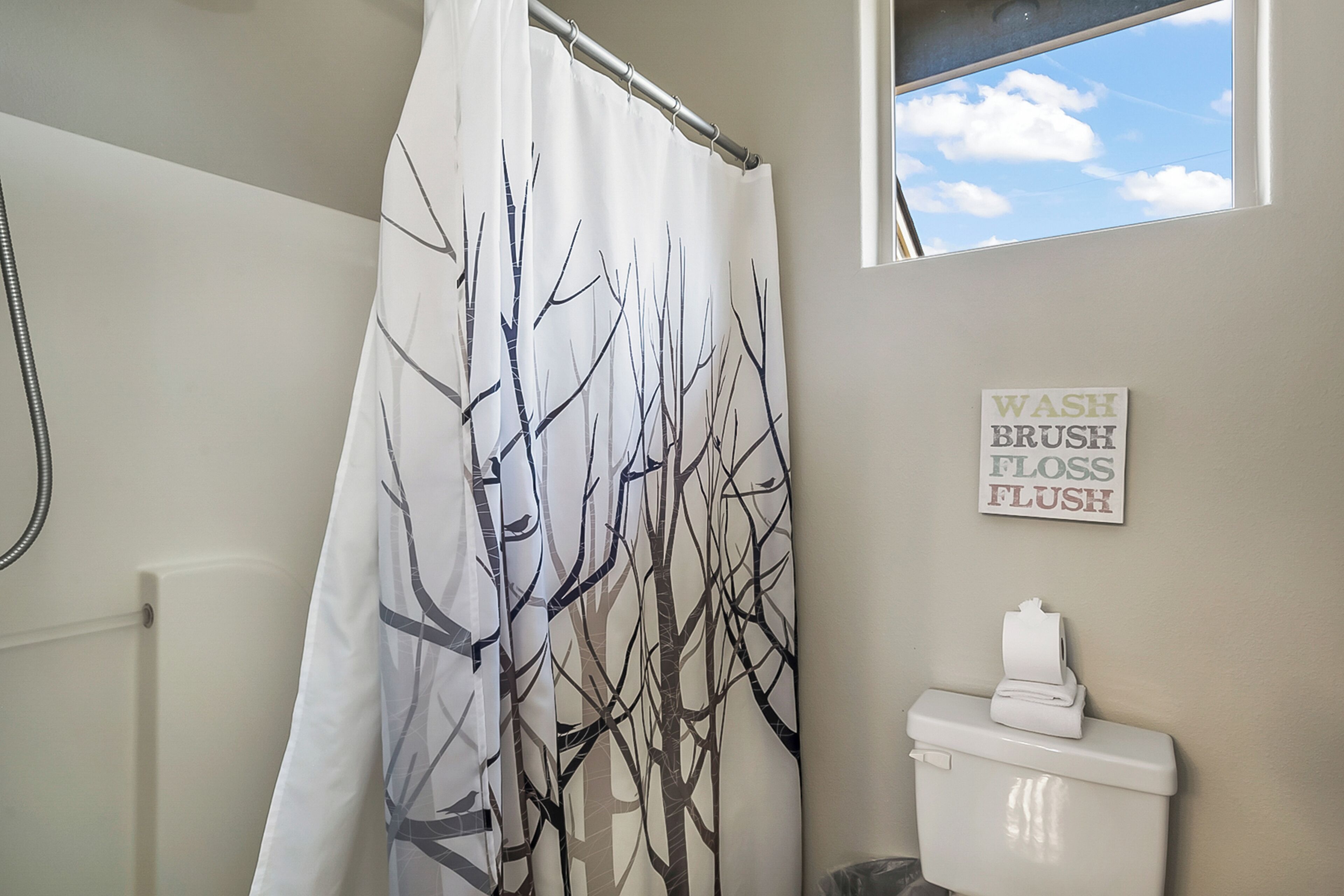 A bathroom with a shower curtain featuring a tree pattern, a toilet, a roll of toilet paper, and a sign on the wall that reads 'WASH BRUSH FLOSS FLUSH'.