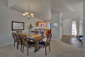 A dining area with a wooden table and chairs, a chandelier, and a kitchen in the background.
