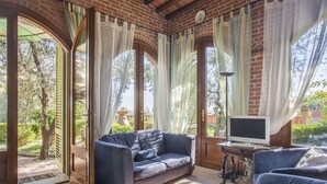 A cozy room with a brick ceiling, large windows with sheer curtains, a blue sofa, a TV on a wooden table, and a view of greenery outside. - Cosy apartment for 4 guests with hot tub, WIFI, pool, TV, patio and pets allowed (Montecatini Terme)