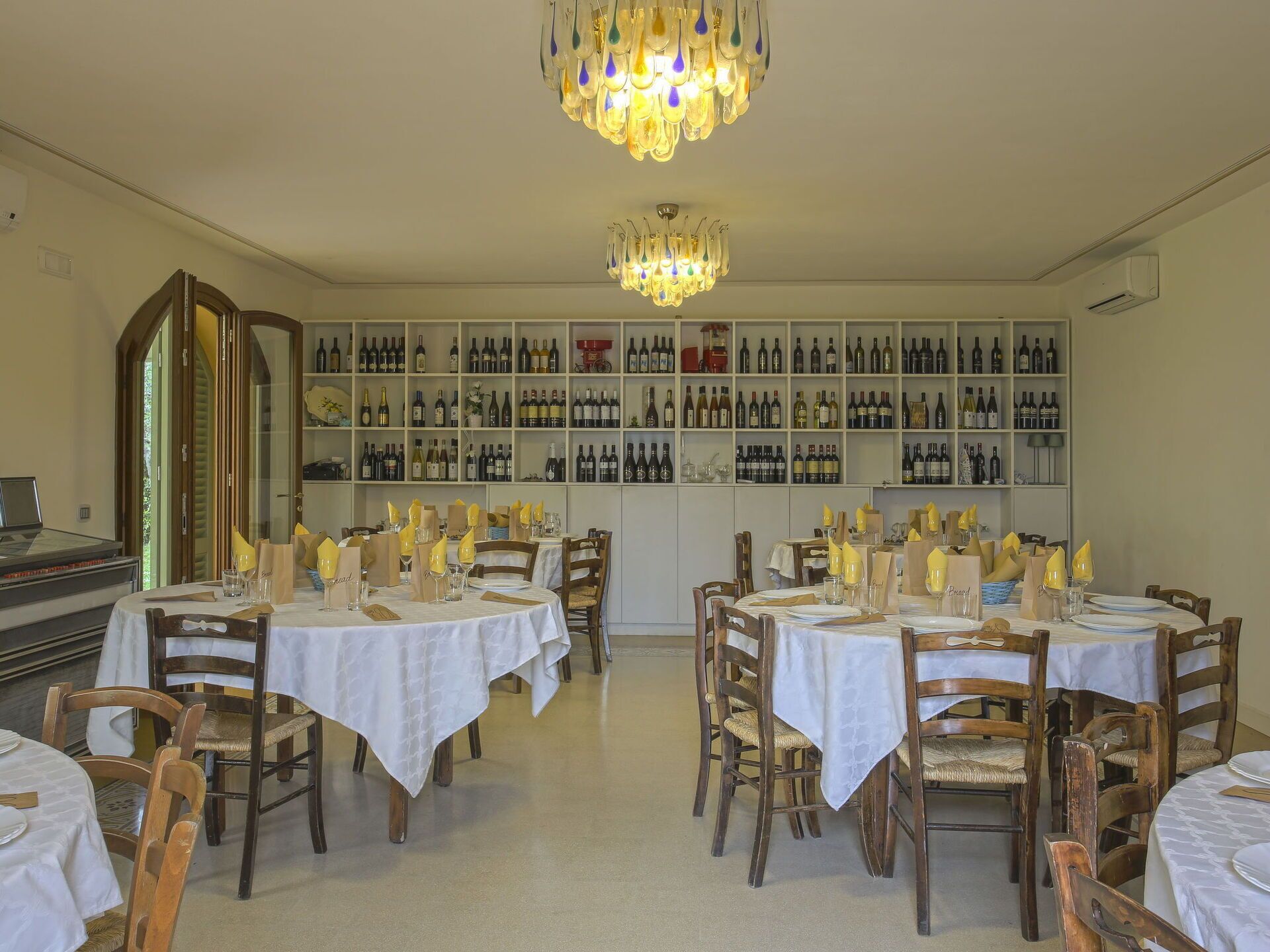 Un comedor con varias mesas, cada una con mantel blanco y sillas de madera. Se ve una bodega grande con numerosas botellas al fondo.