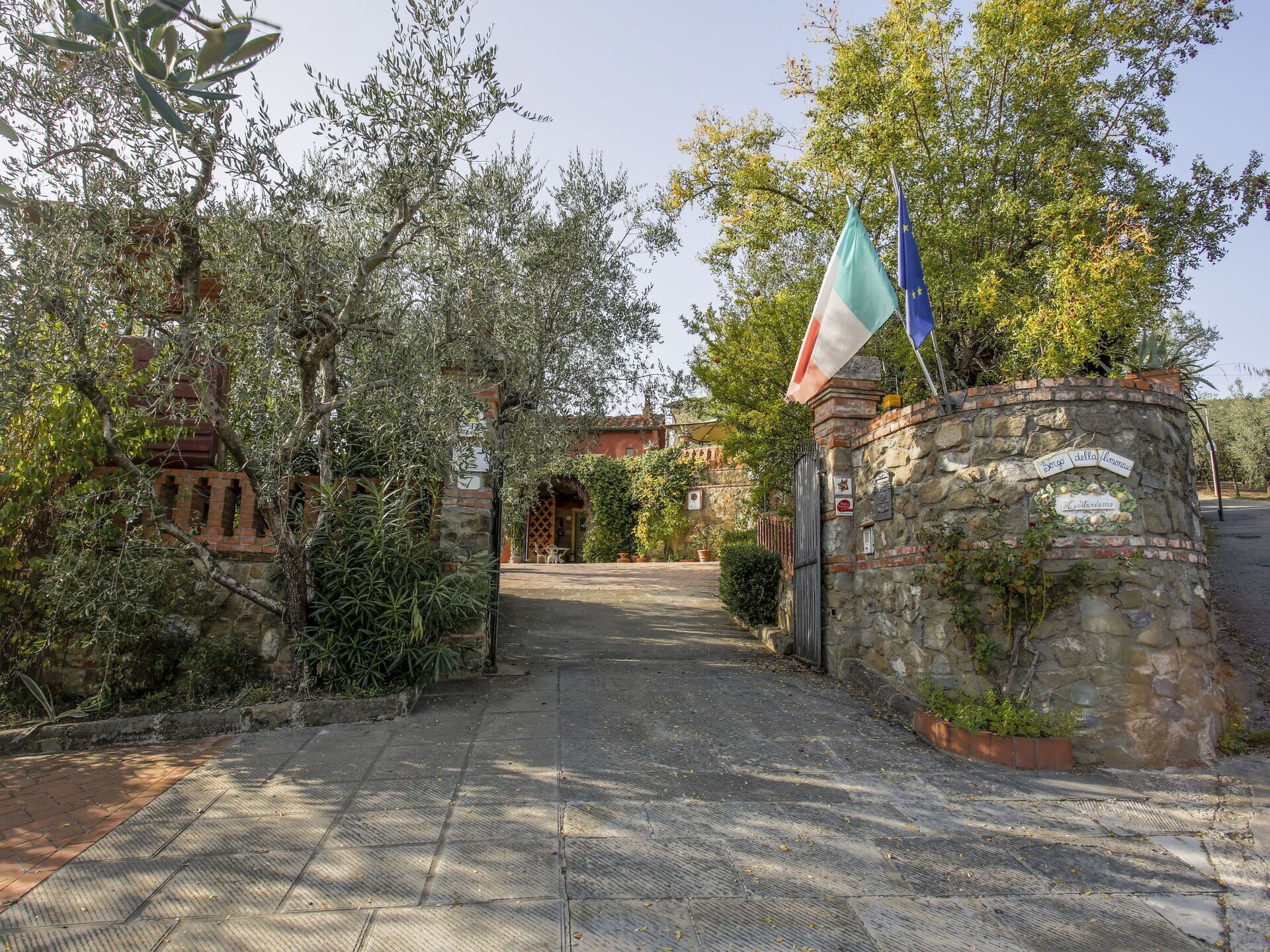 A stone entrance gate with flags, a paved path leading to a building, and lush greenery.