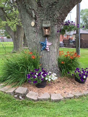A serene tree stands tall, adorned with a birdhouse and star-shaped ornament, surrounded by vibrant flowers and a lush lawn.
