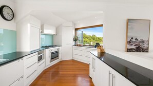 A modern kitchen with white cabinetry, a black countertop, a stove, an oven, and a window with a view of greenery. - The Deck House - sleeps 8 (SMITHS BEACH)