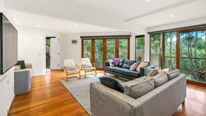 A modern living room with a grey sofa, two armchairs, a TV, and large windows offering a view of trees. - The Deck House - sleeps 8 (SMITHS BEACH)