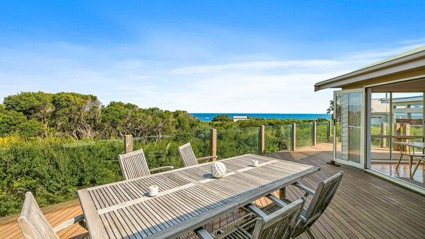 A wooden deck with a table and chairs, overlooking a coastal landscape with trees and the sea. - The Deck House - sleeps 8 (SMITHS BEACH)
