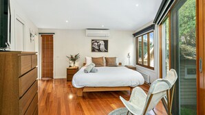 A bedroom with a large bed, wooden flooring, a nightstand with a lamp, a wall-mounted air conditioner, and a window with a view of greenery. - The Deck House - sleeps 8 (SMITHS BEACH)