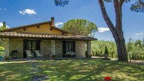 Une maison en pierre avec un toit en tuiles, un patio couvert avec des meubles d’extérieur et une pelouse bien entretenue agrémentée de fleurs rouges.