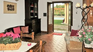 Un salon chaleureux avec un plancher en bois, un buffet en bois foncé, un canapé avec des coussins, une table à manger avec un panier en osier et des fleurs roses, et une vue sur un jardin par une porte ouverte.