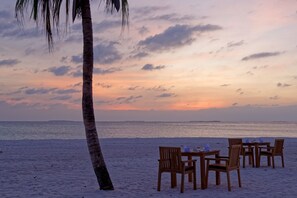 Ein Strand mit Holztischen und -stühlen, Palmen und einem Sonnenuntergang über dem Meer.