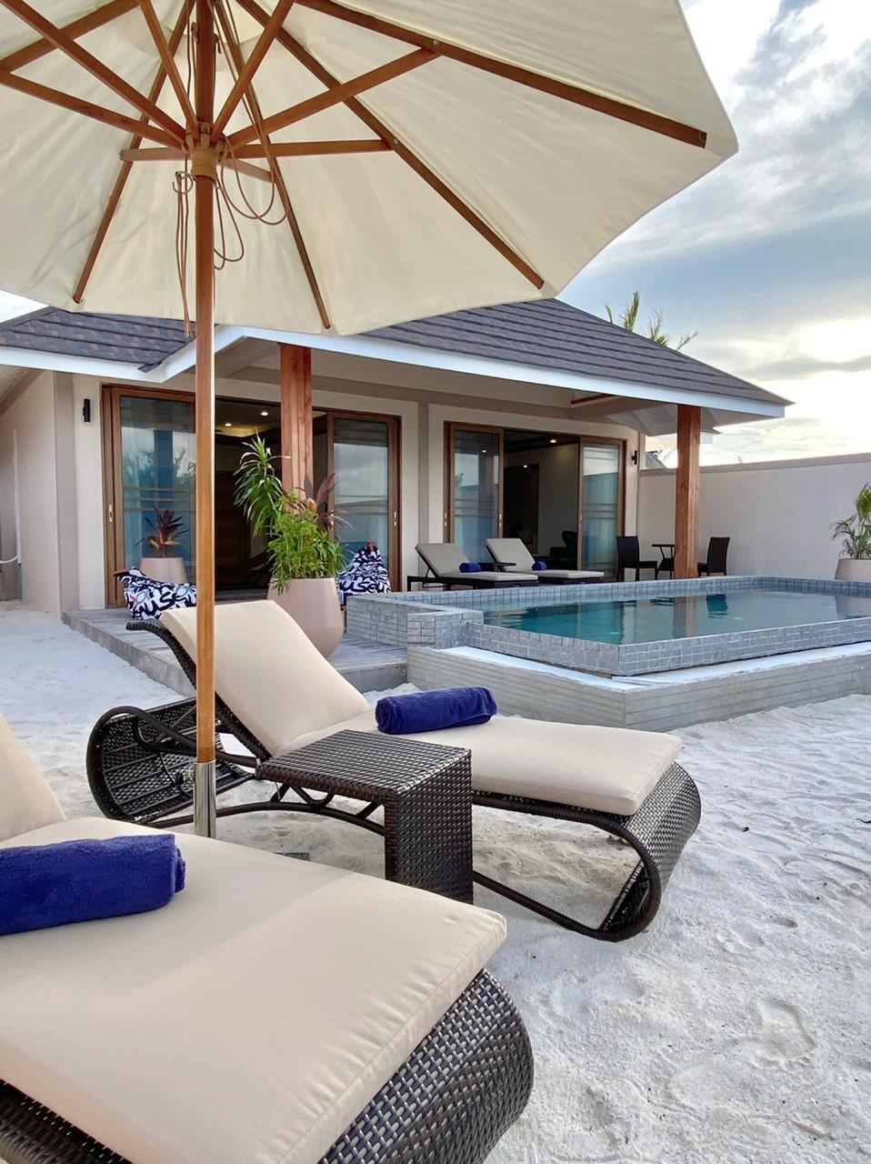 A poolside area with lounge chairs and an umbrella, a pool, and a building with large windows and doors.