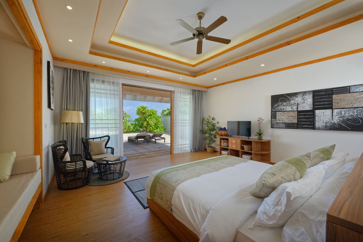 A modern hotel room with a large bed, a ceiling fan, a TV, and a balcony with outdoor seating.