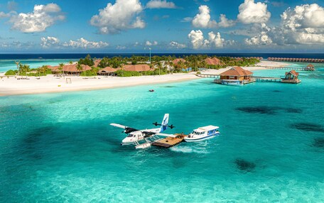 A seaplane docked at a tropical resort with clear turquoise waters, overwater bungalows, and a beach in the background.