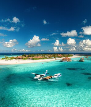 A seaplane docked at a tropical resort with clear turquoise waters, overwater bungalows, and a beach in the background. - Brennia Kottefaru (Kottefaru)