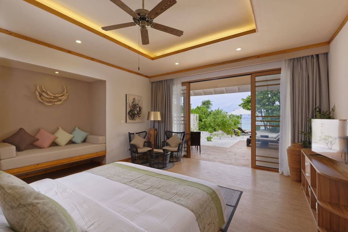 A spacious bedroom with a large bed, a ceiling fan, a sofa with cushions, a dining area with chairs, and a view of the outdoors.