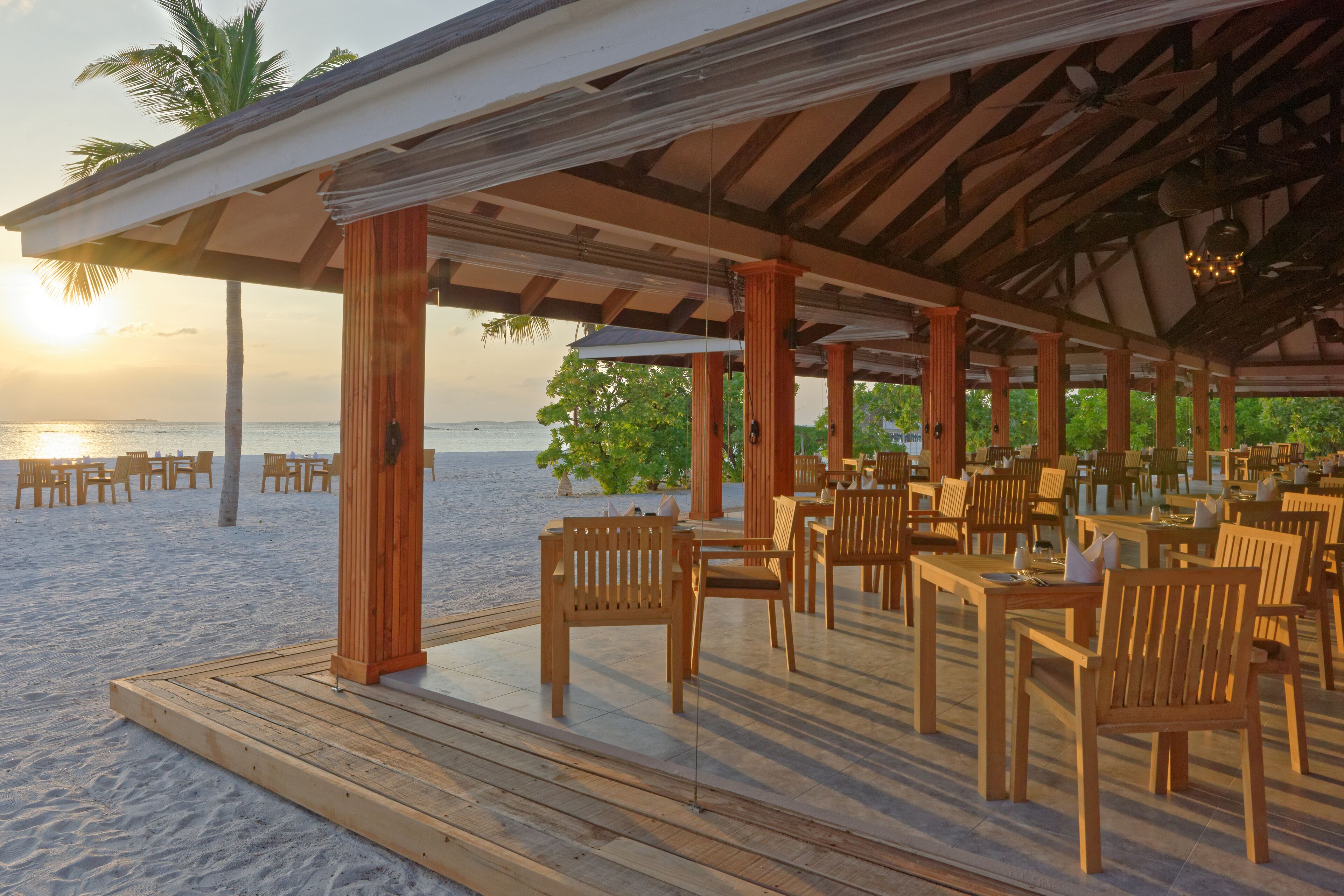 An outdoor restaurant with wooden tables and chairs, a covered patio, and a view of the beach and ocean.