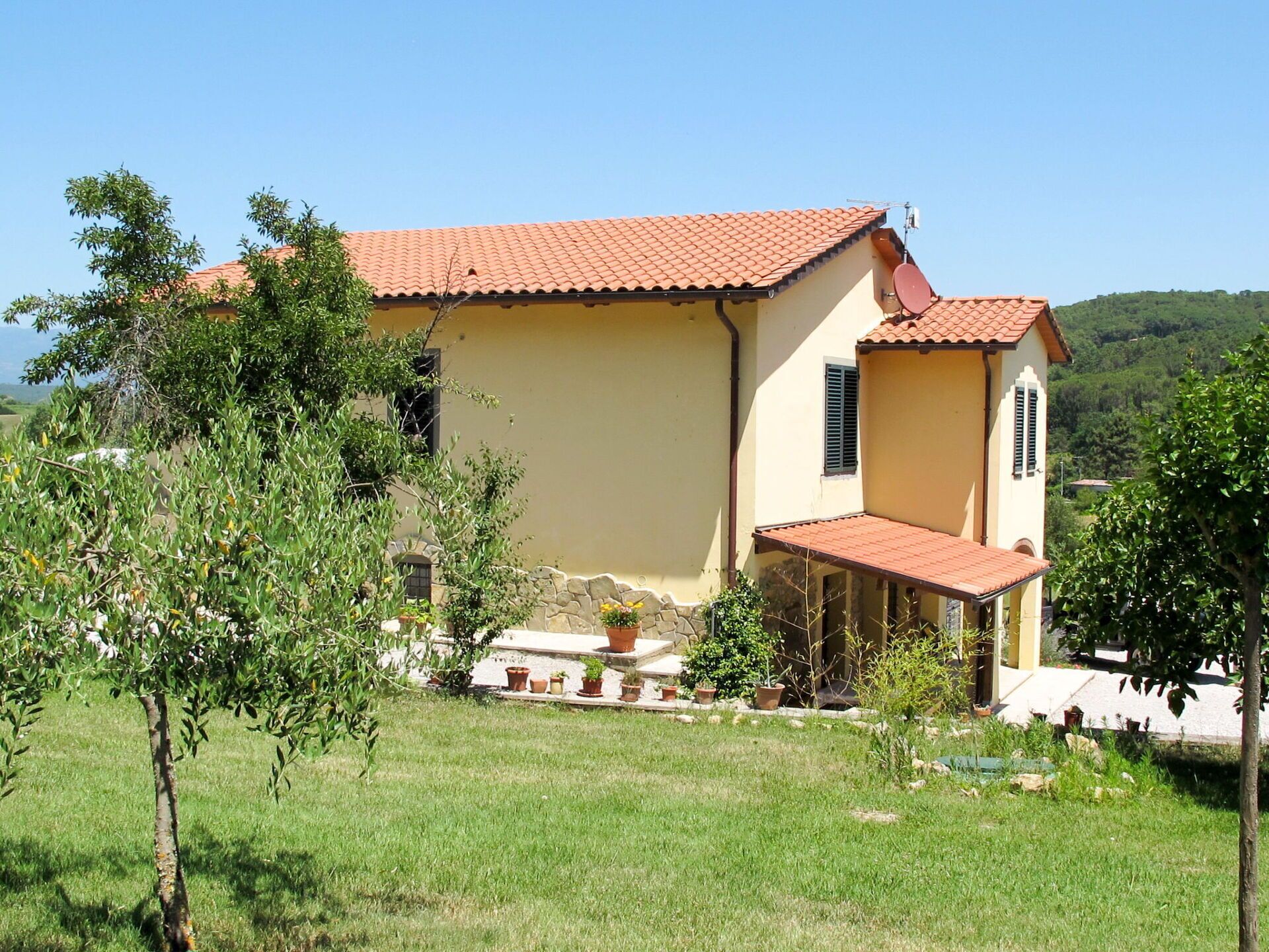 A two-story house with a red-tiled roof, a stone wall, and a small garden area with potted plants.