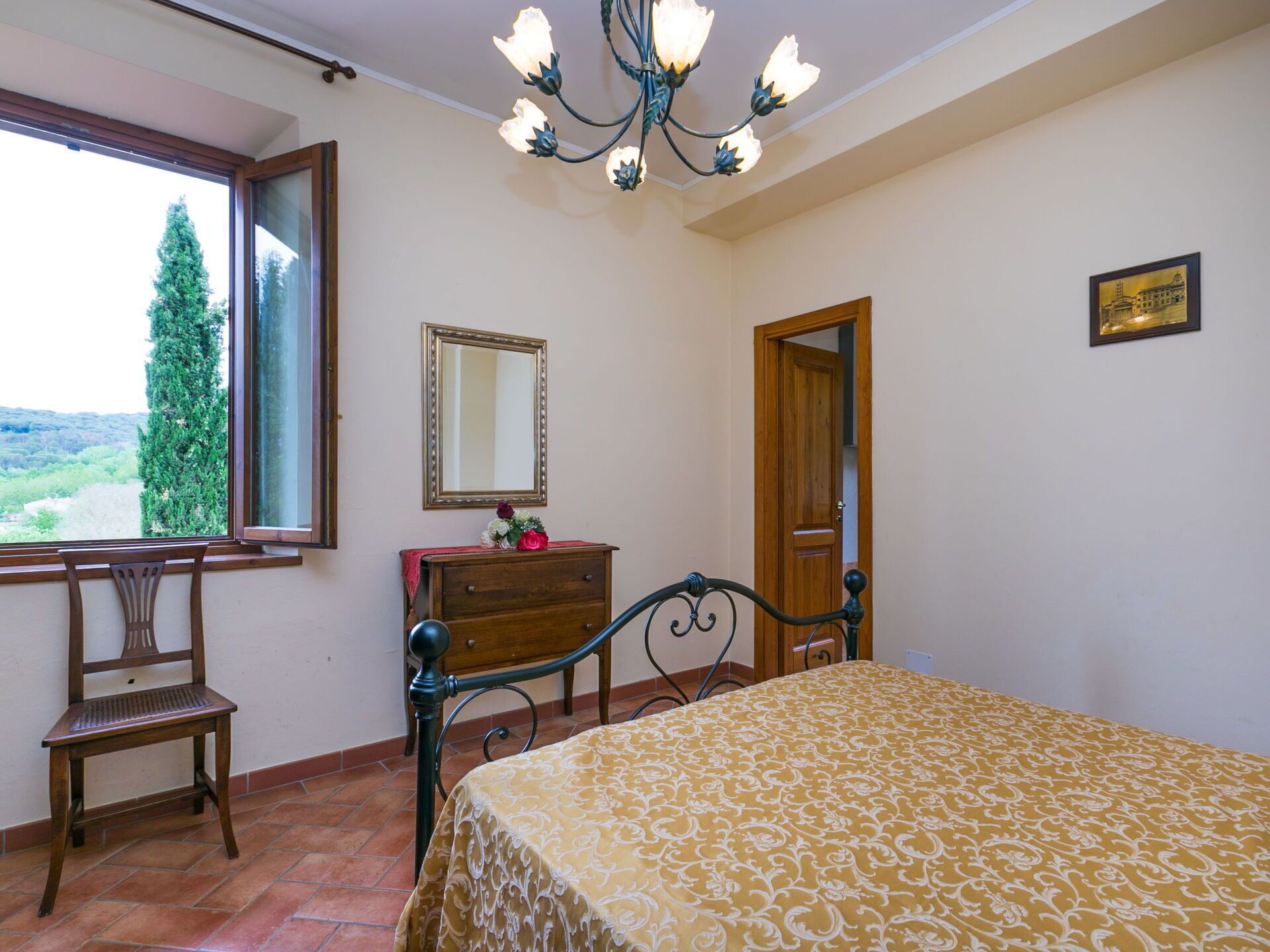 Un dormitorio con cama, una silla de madera, cómoda, espejo y ventana con vista a vegetación.