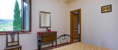 Un dormitorio con cama, una silla de madera, cómoda, espejo y ventana con vista a vegetación.
