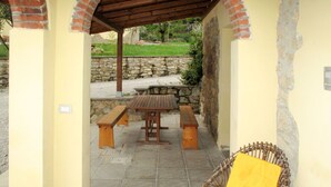 A covered patio area with a stone wall, a wooden table with benches, and a wicker chair with a yellow cushion. - Cosy apartment for 8 people with pool, WIFI, TV, terrace, pets allowed and panoramic view (Pergine Valdarno)
