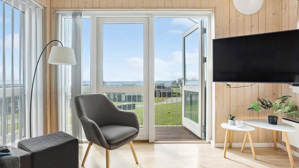 A modern living room with a grey armchair, a small white table with a plant, a mounted TV, and a balcony with a view of the sea. - Pet friendly home in Faaborg with sauna (Faaborg)