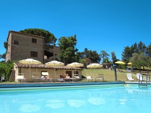 Un espace piscine avec des chaises longues et des parasols, un bâtiment doté d’un toit en tuiles et des arbres en arrière-plan.
