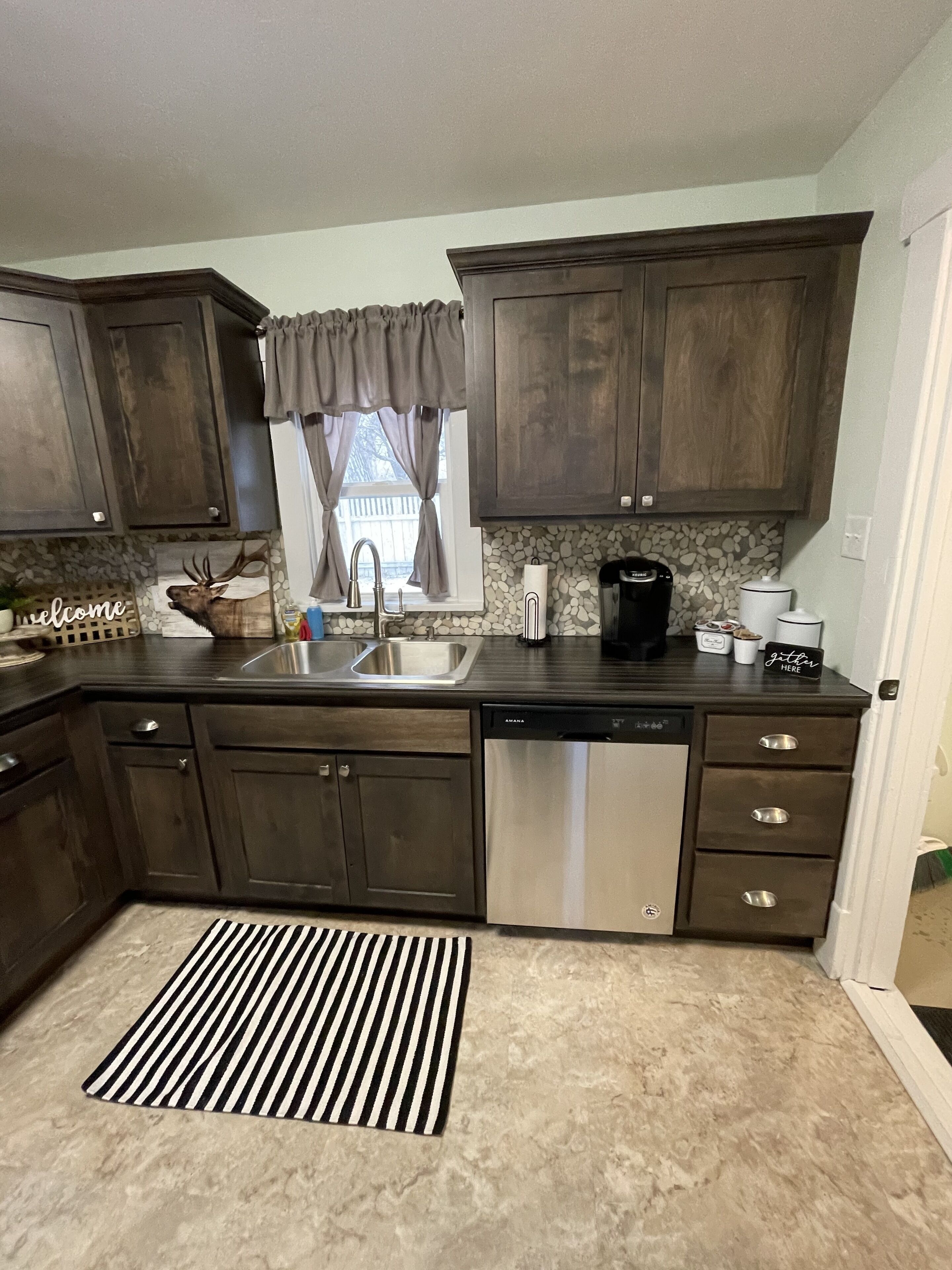 A kitchen with dark wooden cabinets, a stainless steel dishwasher, a coffee maker, and a striped rug.