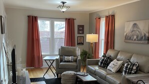 Un salon comprenant un canapĂ©, un fauteuil, une table basse et un tĂ©lĂ©viseur posĂ© sur un meuble. On y trouve Ă©galement un ventilateur de plafond, une lampe et une fenĂȘtre avec des rideaux.