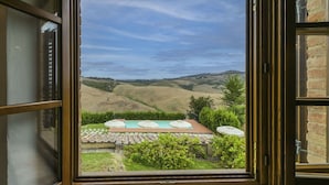 Vista attraverso una finestra aperta su una zona piscina con tavolo e ombrelloni, immersa nel verde e colline.
