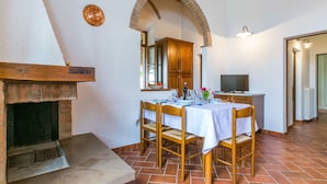 Un comedor con chimenea de ladrillo, una mesa de madera y sillas. Se observa una zona de cocina con armarios y un televisor.