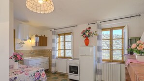 Una cuina amb terra de rajola, nevera, rentapelats, armaris i una finestra amb cortines.