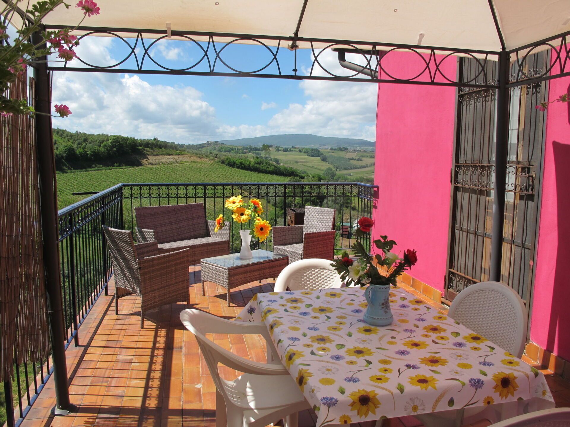 A balcony with wicker furniture, a table with a floral tablecloth, and a vase of flowers.
