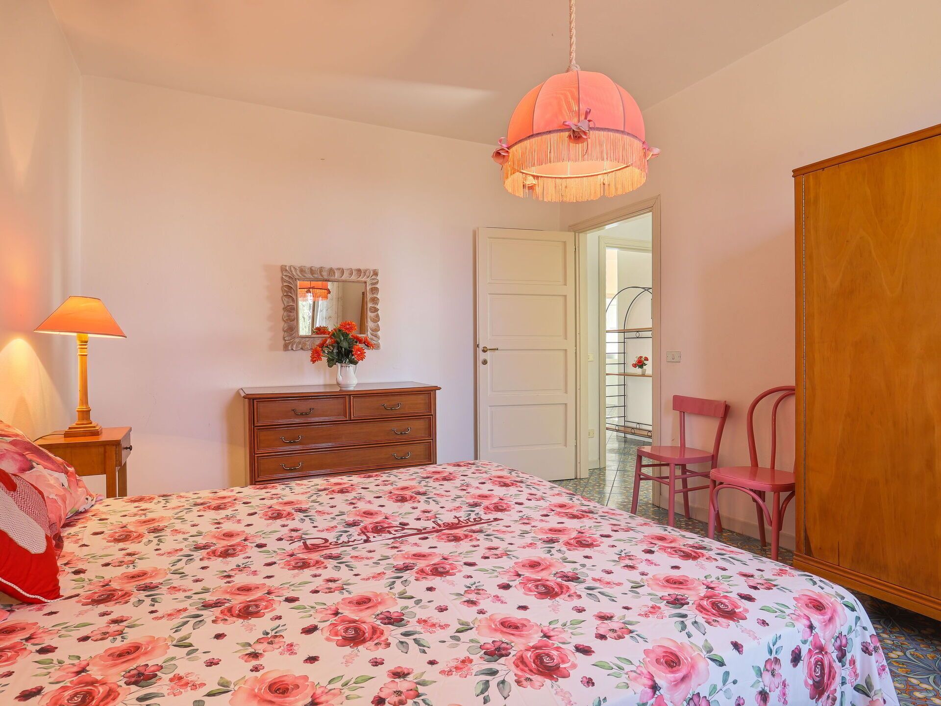 A bedroom with a floral bedspread, a wooden dresser, a mirror, and a pink hanging light fixture.