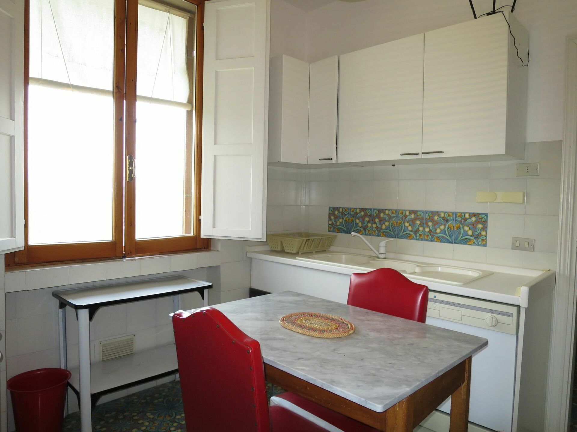 A compact kitchen with white cabinets, a tiled backsplash, a dining table with chairs, and a window with curtains.
