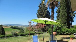 Un patio con due poltrone blu sotto un ombrellone verde, un muro in mattoni e vista su un vigneto e delle colline sullo sfondo.
