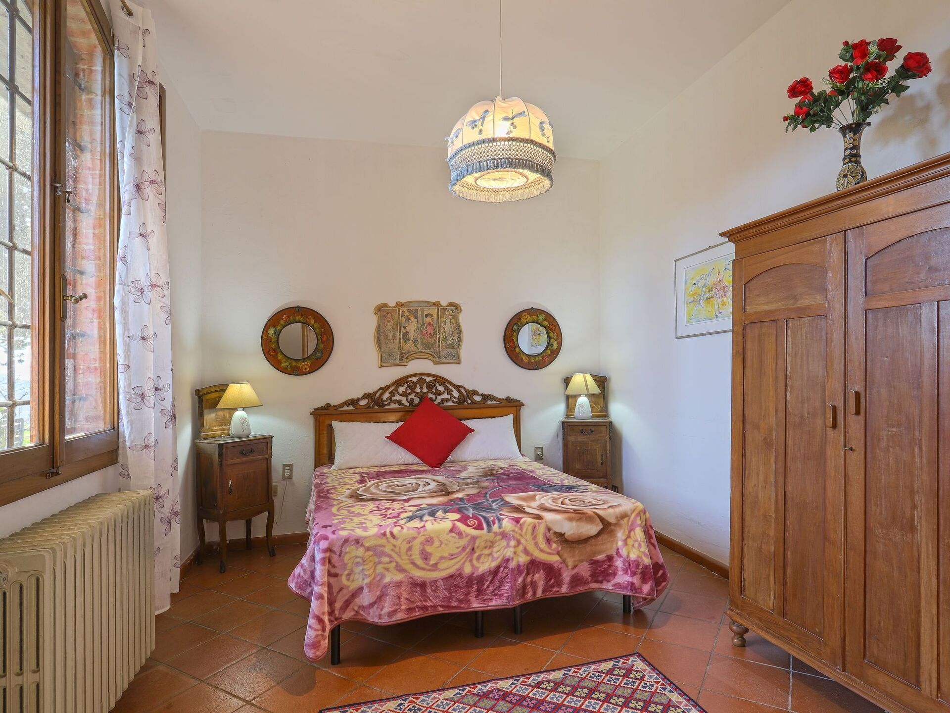 Une chambre à coucher comprenant un lit, une armoire en bois, un radiateur, une fenêtre avec des rideaux et un luminaire suspendu.
