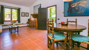A dining area with a round wooden table, chairs, and a bench. There is a painting on the wall and a window with curtains. - Cosy apartment in villa for 2 guests with pool, WIFI, TV and terrace (Colle Di Val D'elsa)