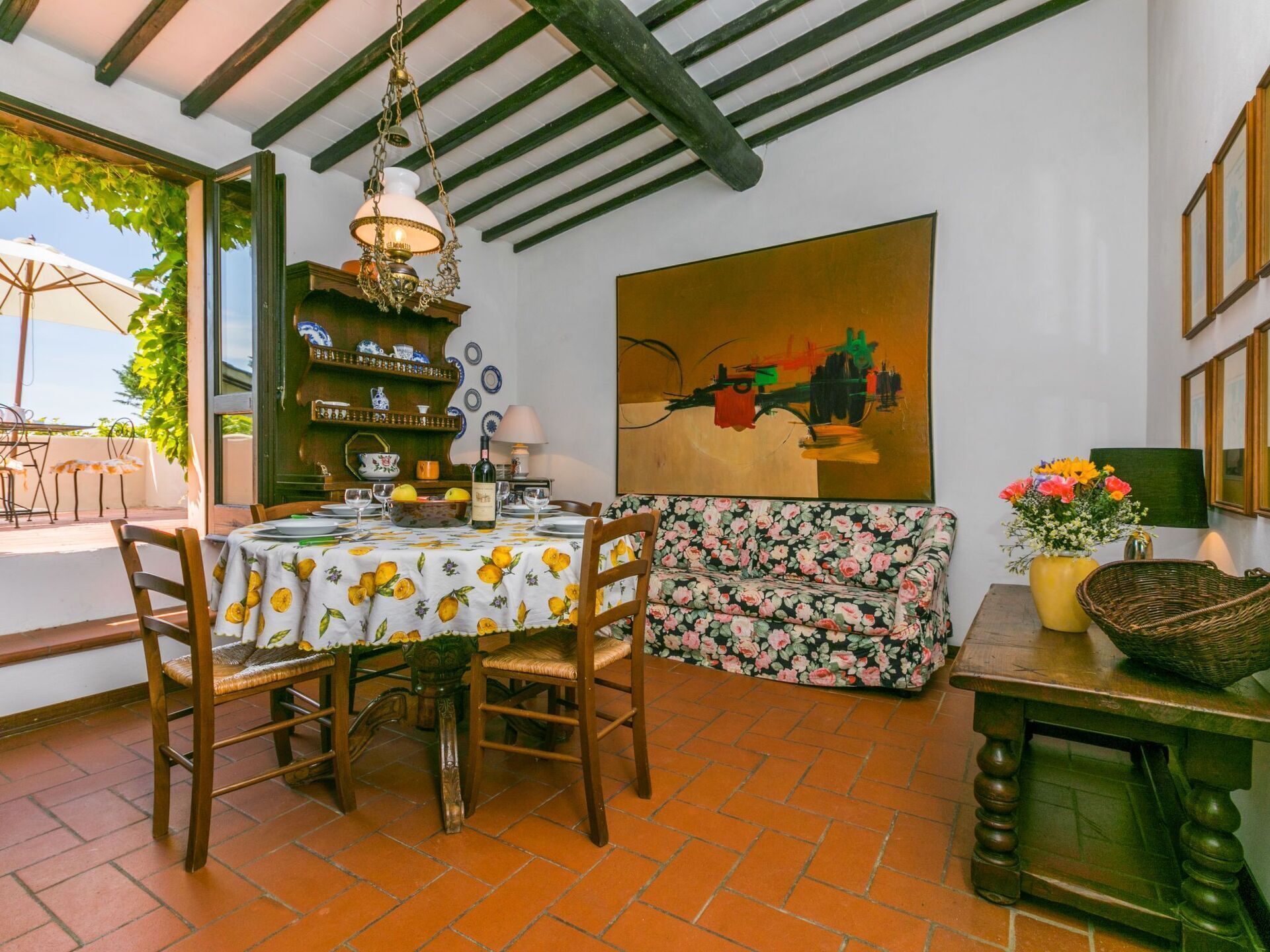 A dining area with a table set for two, a floral sofa, and a painting on the wall.
