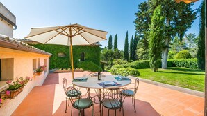 A patio with a round table and chairs, an umbrella, and a garden with trees and shrubs.