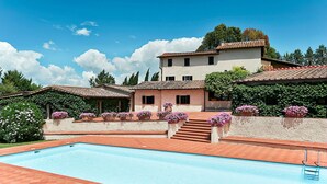 A pool area with a clear blue pool, terracotta tiled deck, and a building with a tiled roof and stucco walls.