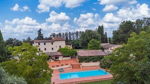 A pool area with a rectangular pool, surrounded by a red-tiled deck and green lawn. There are lounge chairs and a small staircase leading to a building with a tiled roof.