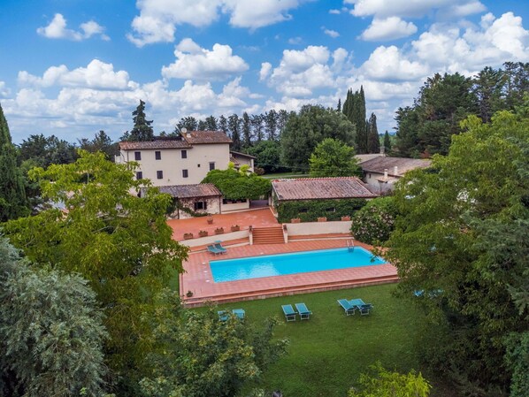 Un espace piscine doté d’une piscine rectangulaire, entourée d’une terrasse carrelée de tuiles rouges et d’une pelouse verdoyante. On y trouve des chaises longues et un petit escalier menant à un bâtiment dont le toit est également recouvert de tuiles.