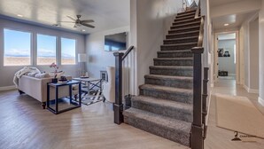 A spacious living room with a large window, a staircase, and a flat-screen TV. - Parking galore, quiet & bonus- electrical RV hookup! (Saint George)