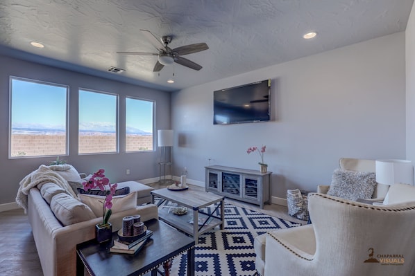 A spacious living room with a ceiling fan, large windows with a mountain view, a flat-screen TV, and a cozy seating area with a patterned rug.
