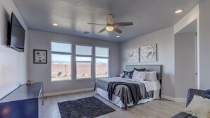 A bedroom with a bed, a ceiling fan, a TV, and large windows with a view of the mountains.