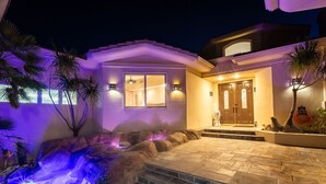 A modern house with a stone patio, a small waterfall feature, and a well-lit entrance. - Hidden Oasis: Pickleball Basketball Waterslide Game Room Gym! ⭐️⭐️⭐️⭐️⭐️ (Palm Desert)