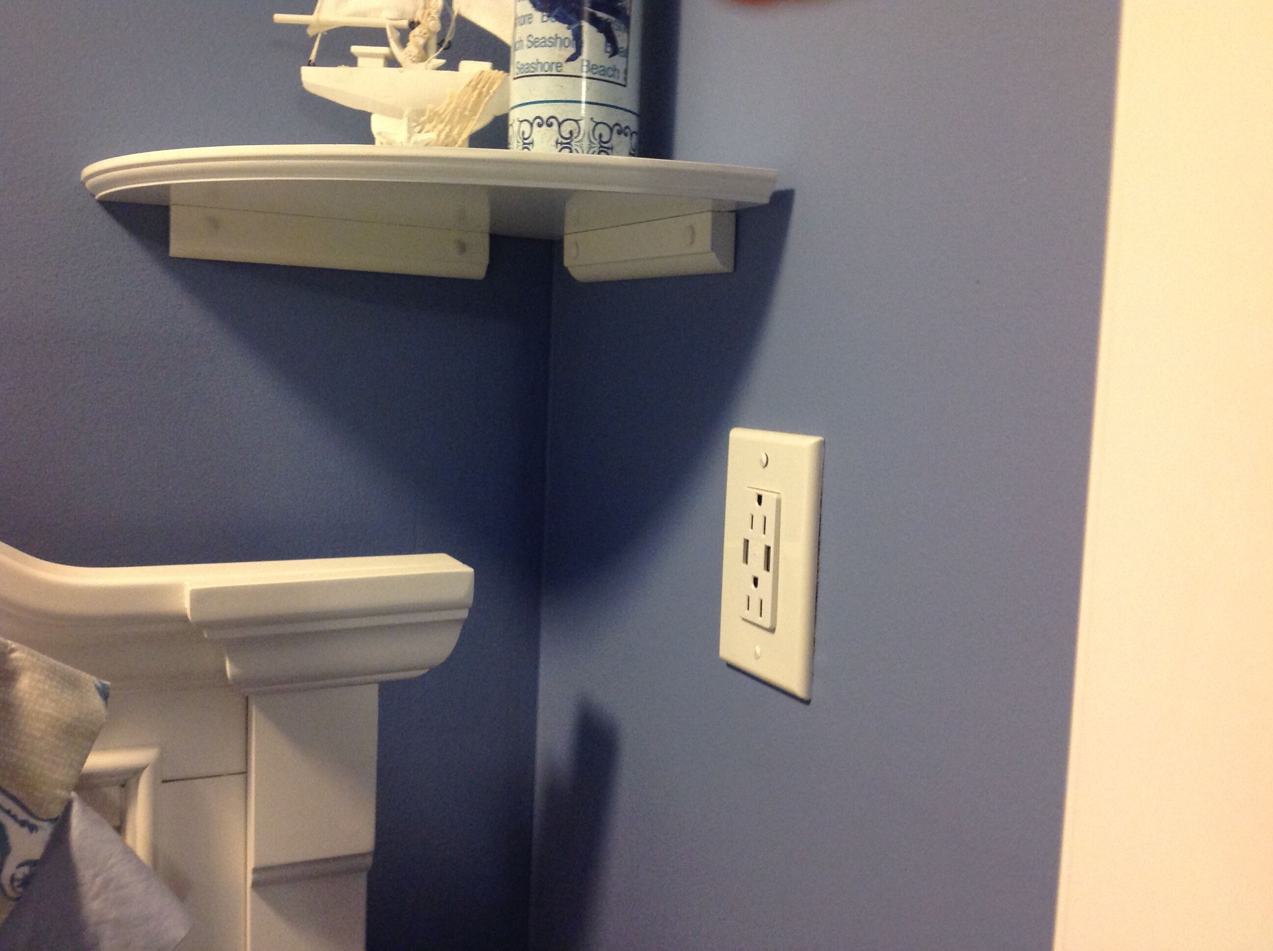 A bathroom with a white toilet, a shelf with a bottle, and a power outlet on the wall.